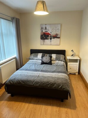 Quarto com cama, mesa de cabeceira, luminária, janela com cortinas e um quadro na parede.