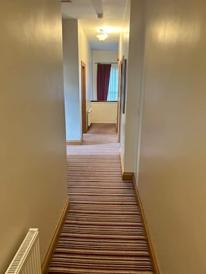 A hotel hallway with a carpeted floor, a radiator, and a window with curtains. - Greencastle – 4-Bed Townhouse Sleeps 8 | Free Parking | Mourne Mountains (Kilkeel)