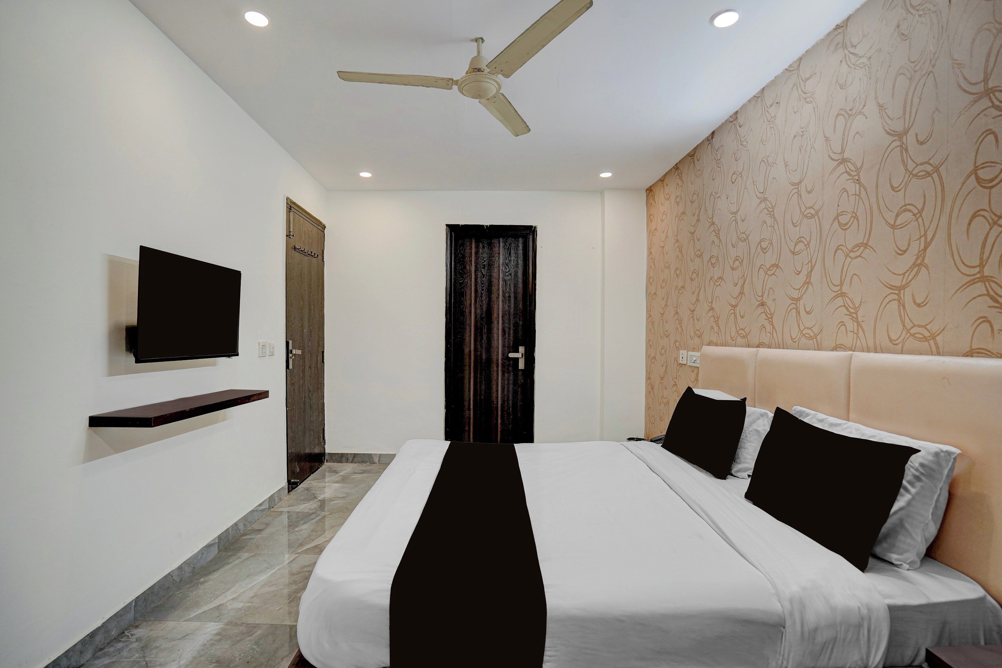 Una habitación de hotel con cama, televisor montado en la pared, ventilador de techo y papel tapiz estampado.