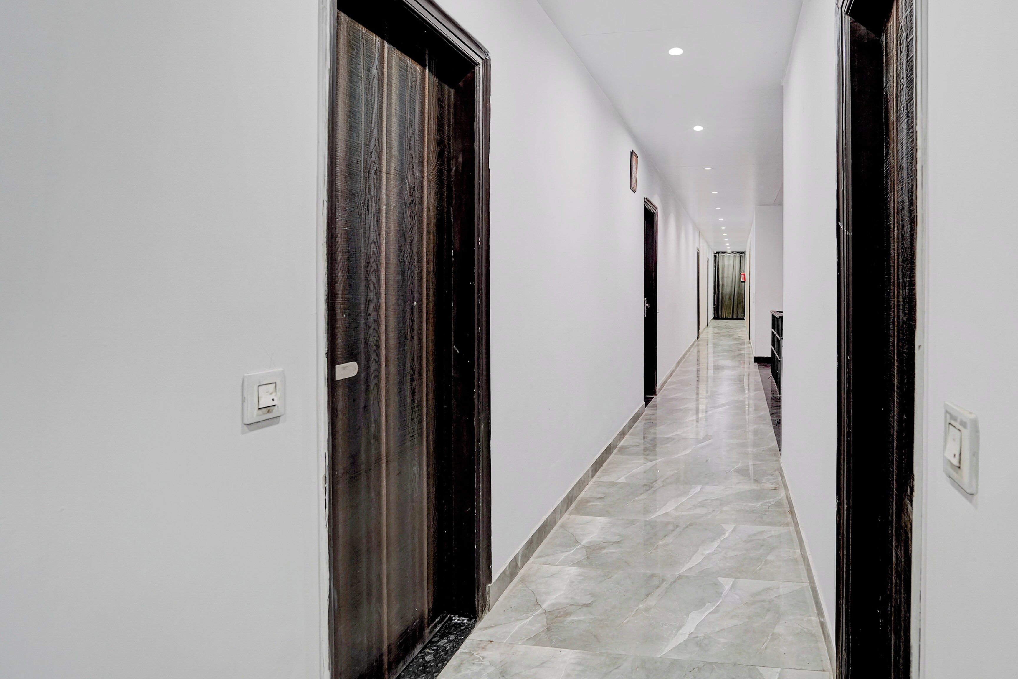 A corridor with white walls and dark wooden doors, leading to a room with a large window.
