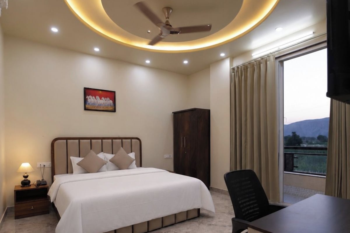 A hotel room with a bed, a desk, a chair, a ceiling fan, and a window with curtains.