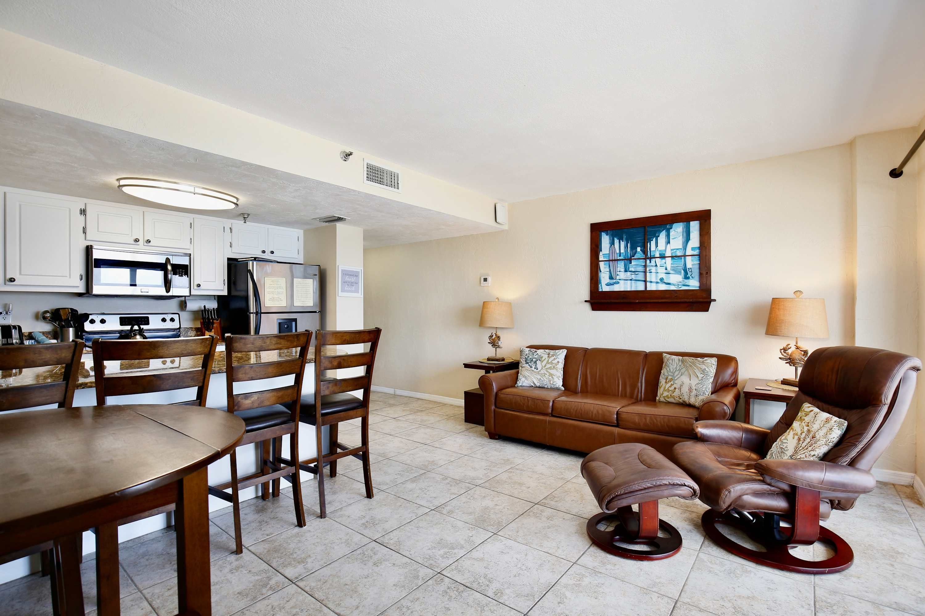 A modern living room with a brown leather sofa, a dining table with chairs, and a kitchen area with white cabinets and stainless steel appliances.