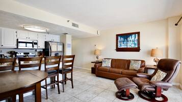 A modern living room with a brown leather sofa, a dining table with chairs, and a kitchen area with white cabinets and stainless steel appliances.