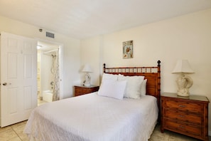 Quarto com cama de madeira, roupa de cama branca, duas mesas de cabeceira com luminárias, uma porta para um banheiro e uma obra de arte na parede.