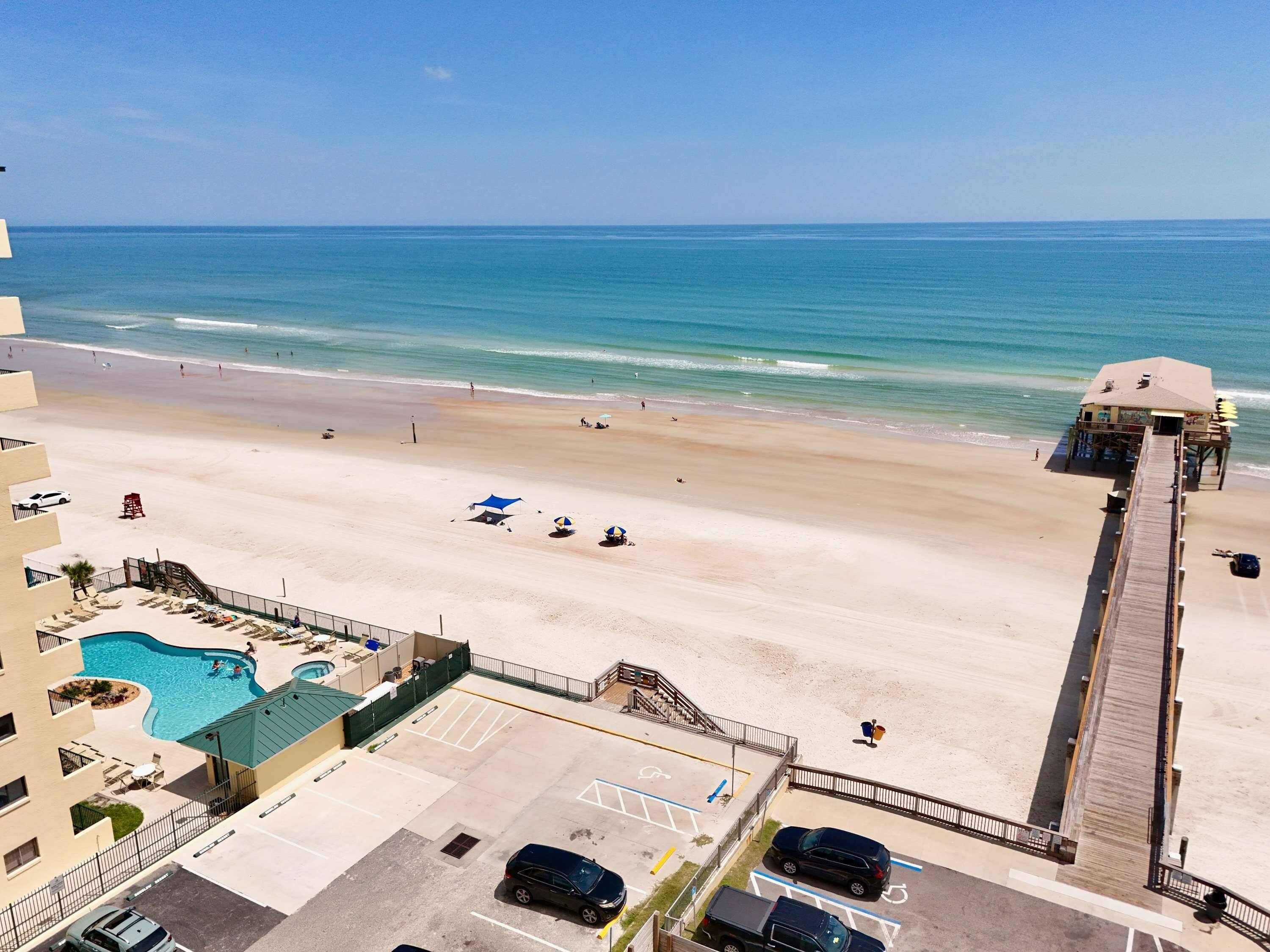 A beach view from a hotel balcony with a pool, beach umbrellas, and a pier.