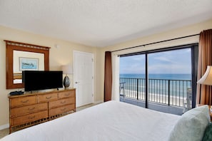 Quarto de hotel com uma cama, televisão sobre uma cômoda de madeira, uma porta e varanda com vista para a praia.