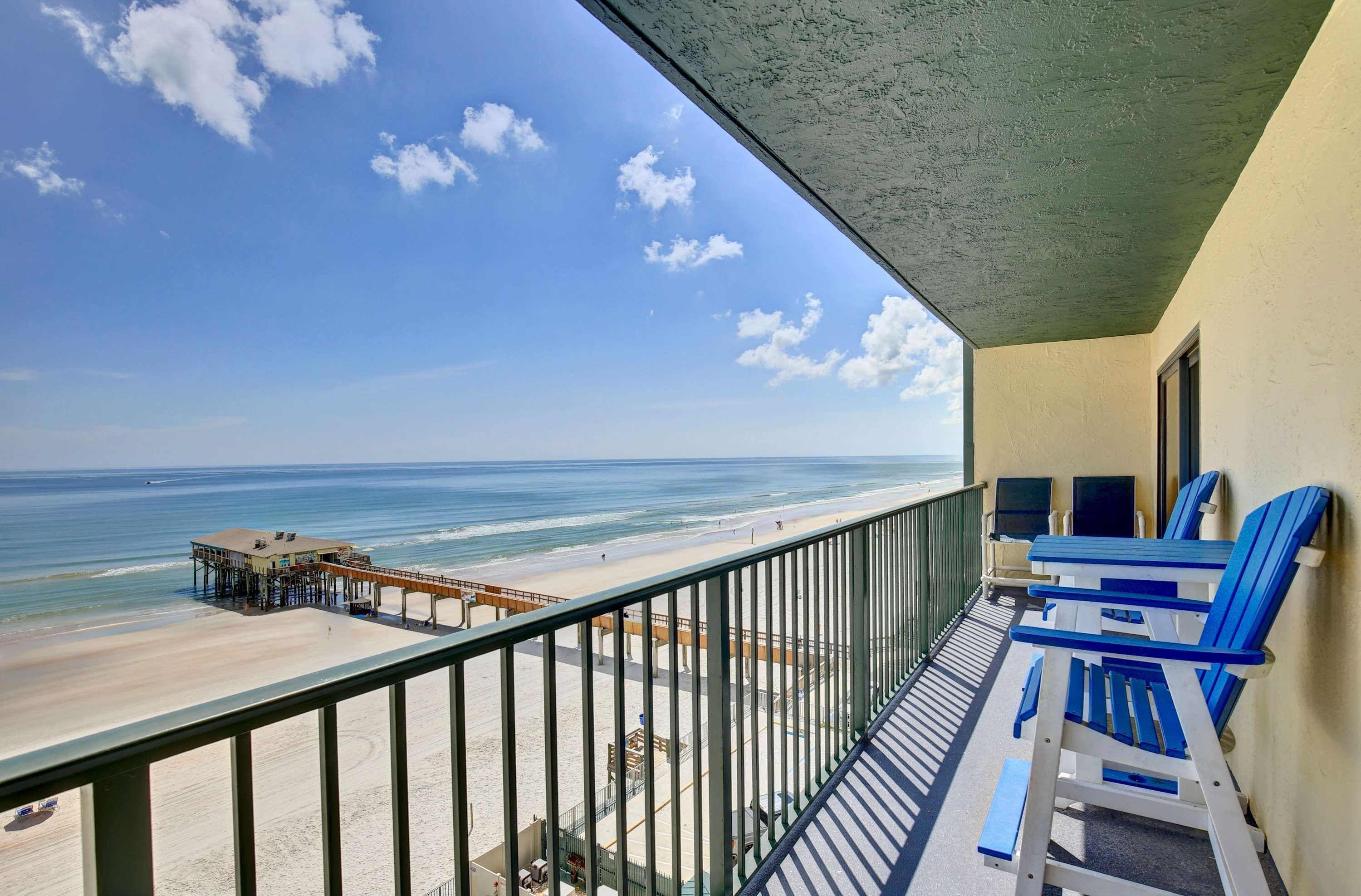 Un balcón con vista a la playa y al océano, que cuenta con una terraza de madera y sillas azules.