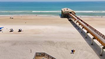 A beach with a wooden pier extending into the ocean, a sandy beach, and a few umbrellas.