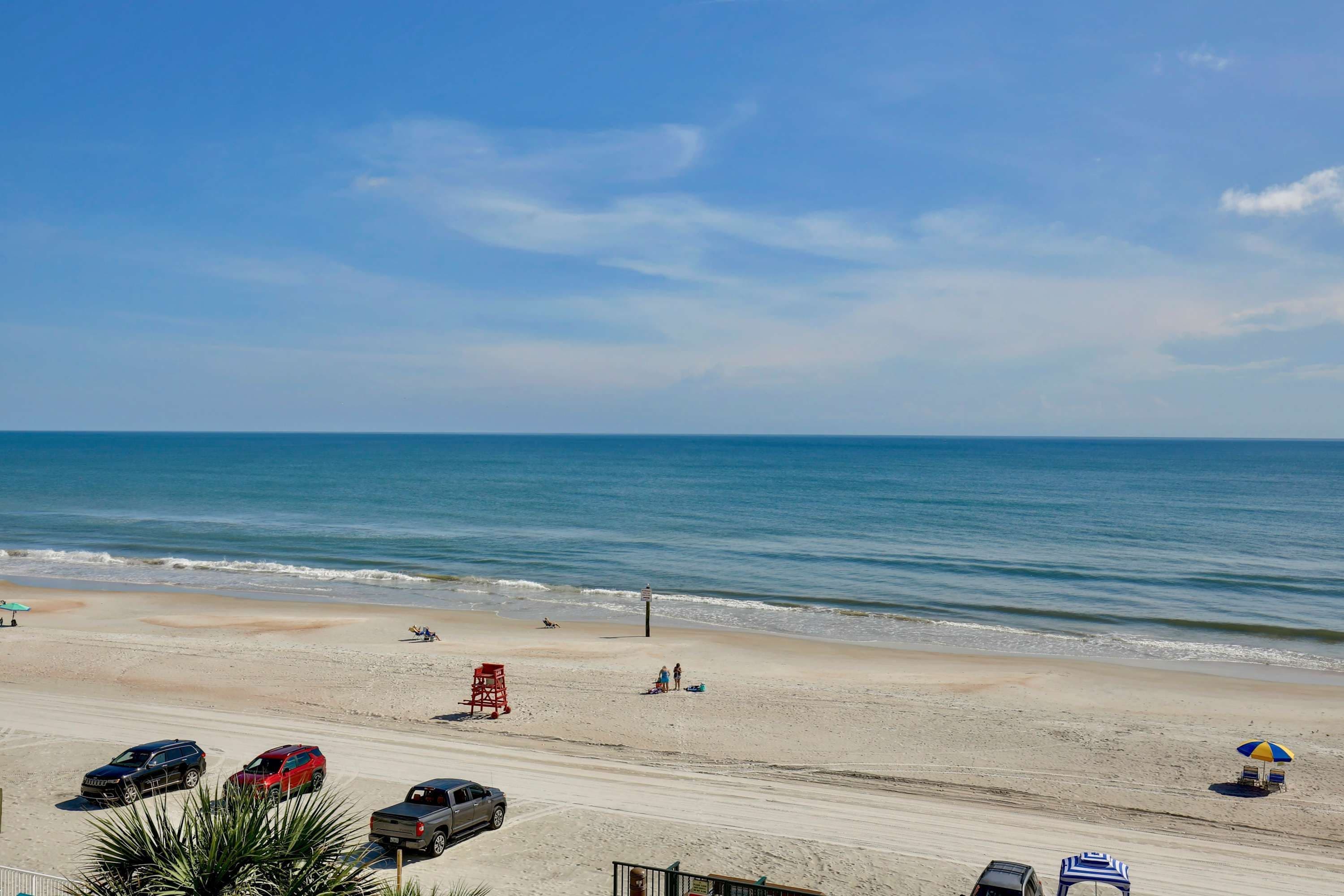 Una playa con cielo despejado, mar tranquilo y varios coches estacionados.