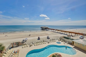 Um resort à beira-mar com piscina, guarda-sóis, espreguiçadeiras e um píer que se estende para o oceano.