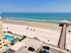 Vista da praia a partir de uma varanda de hotel, com piscina, guarda-sóis e um píer.