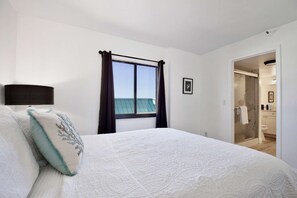 Quarto com cama grande, janela com cortinas, quadro na parede e banheiro visível através de uma porta aberta.