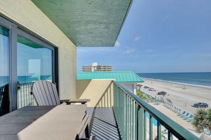 A balcony with a view of the beach and ocean, a table with chairs, and a pool area with lounge chairs. - The Coastal Current at Tropical Suites Sunglow Resort (Daytona Beach Shores)