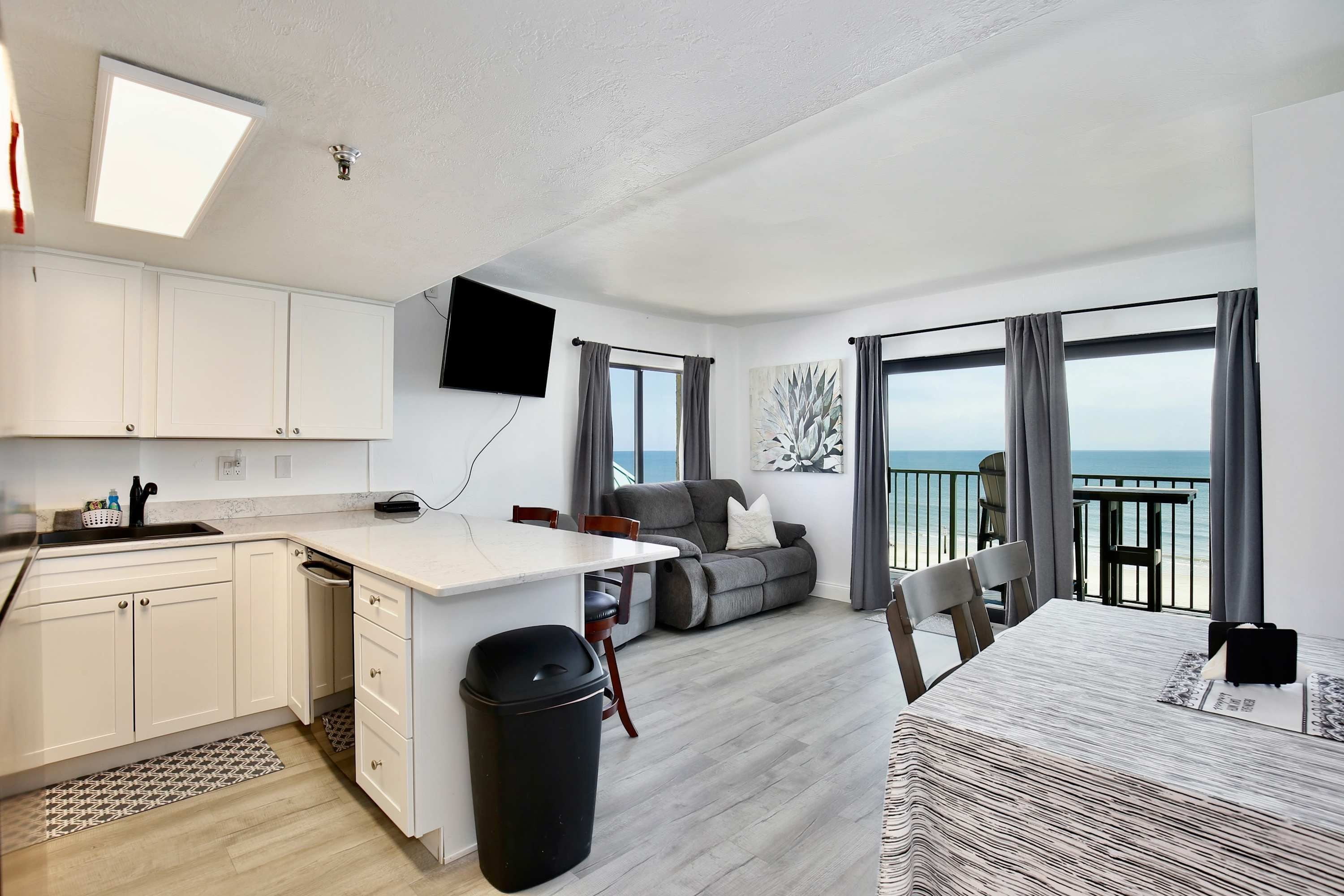 A modern kitchen with white cabinets, a dining area with a table and chairs, a sofa, and a balcony with a view of the sea.