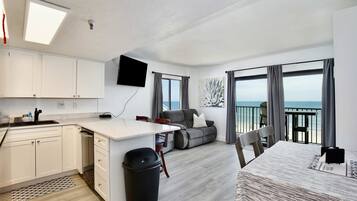 A modern kitchen with white cabinets, a dining area with a table and chairs, a sofa, and a balcony with a view of the sea.