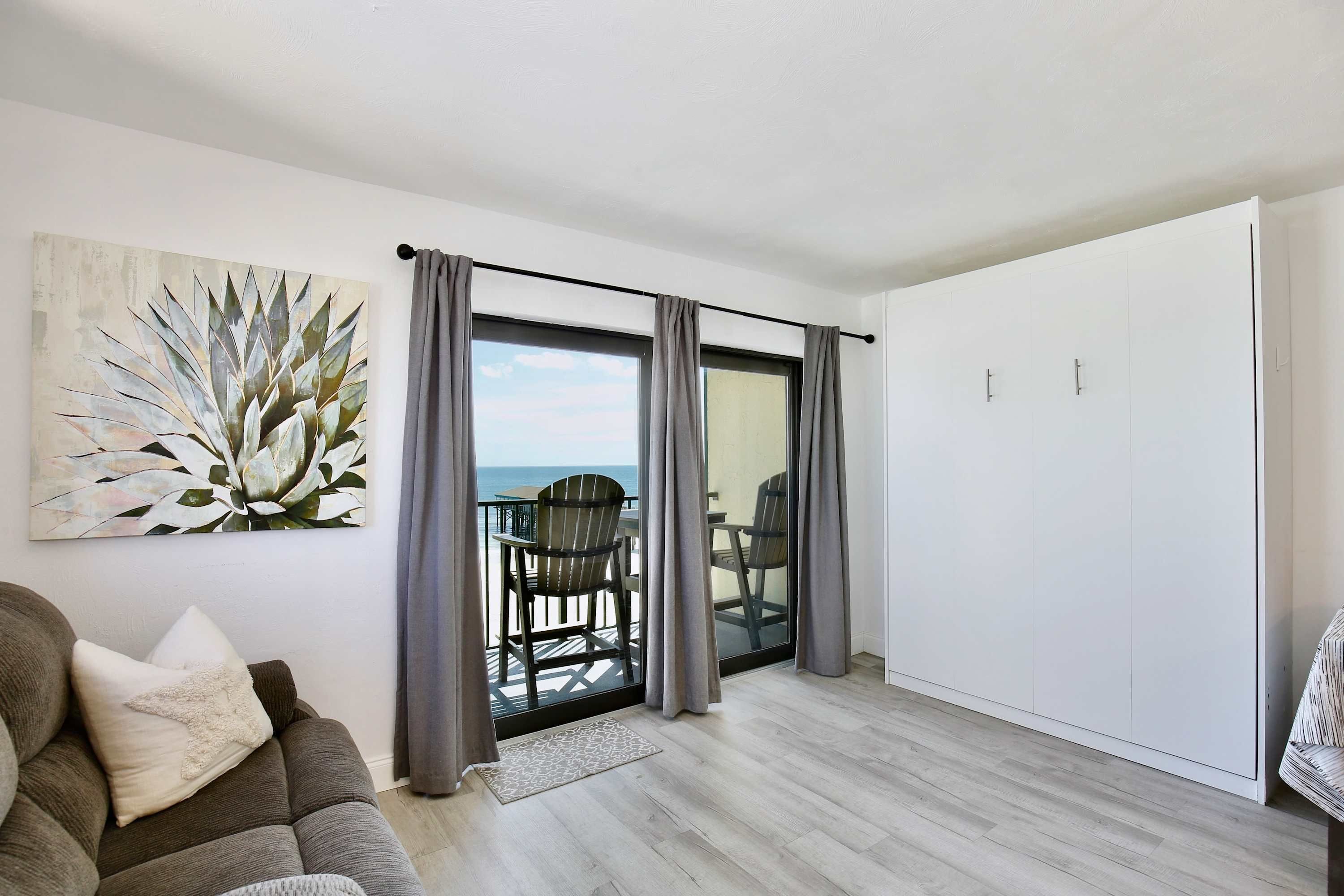A modern living room with a view of the sea, a grey sofa, a painting, and a balcony with chairs.