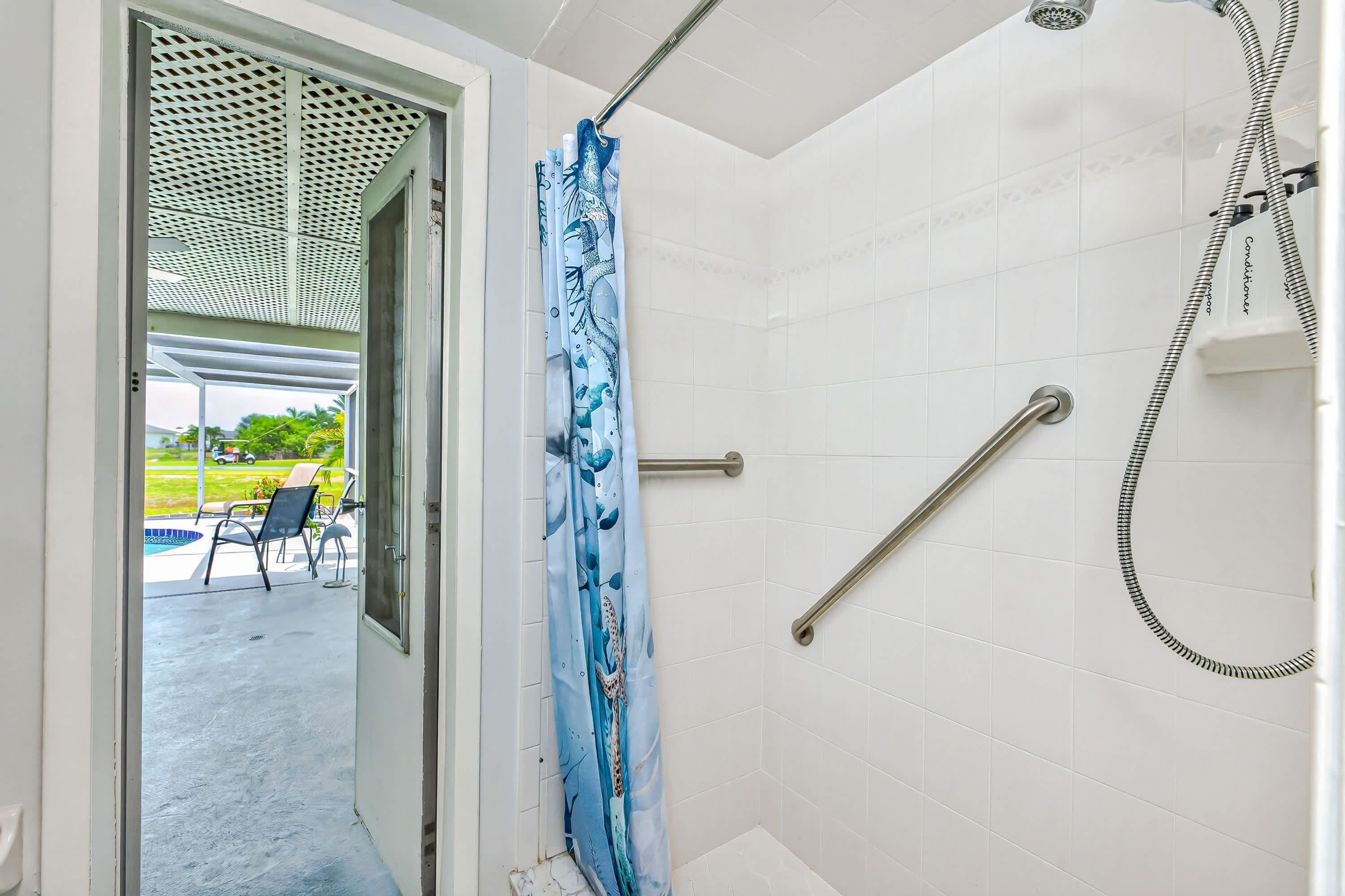 Een badkamer met witte tegels, een douche met handdouche en een glazen deur die leidt naar een buitenruimte met stoelen en een zwembad.