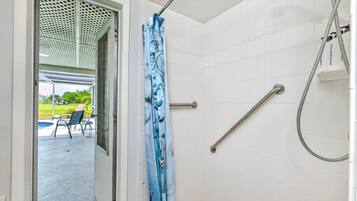 Een badkamer met witte tegels, een douche met handdouche en een glazen deur die leidt naar een buitenruimte met stoelen en een zwembad.