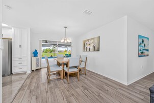 Una cocina moderna con armarios blancos, un comedor con mesa y sillas de madera, y vista a una piscina a través de un ventanal.