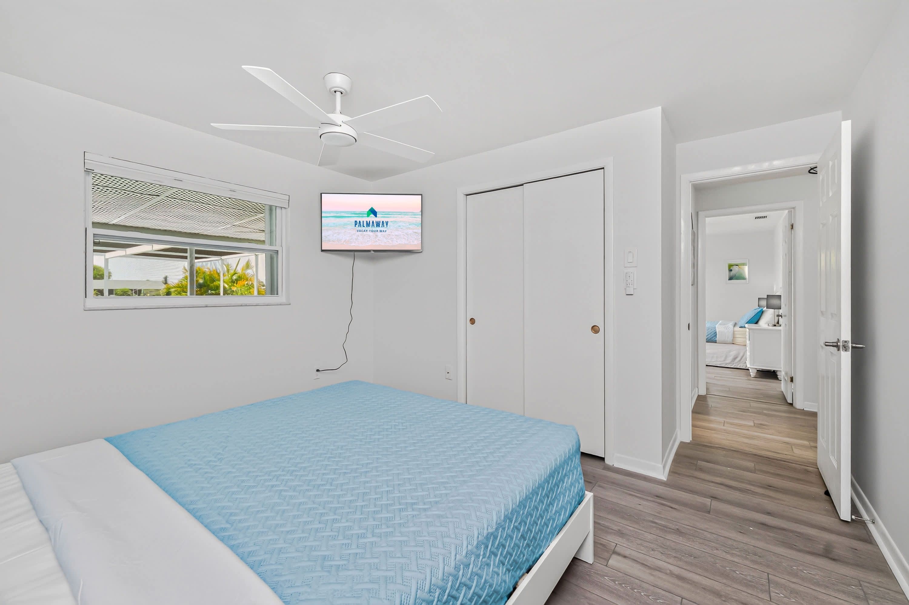 A bedroom with a bed, a ceiling fan, a TV, and a window with a view of greenery.