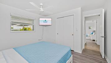 A bedroom with a bed, a ceiling fan, a TV, and a window with a view of greenery.