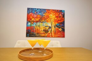 A dining table with a tray holding two glasses of orange juice, a painting of a colorful autumn scene on the wall, and a white chair. - Boutique stay in Athens center (Athina)