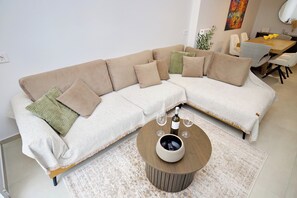A modern living room with a beige sectional sofa, a round coffee table with a bottle and glasses, and a dining area in the background. - Boutique stay in Athens center (Athina)