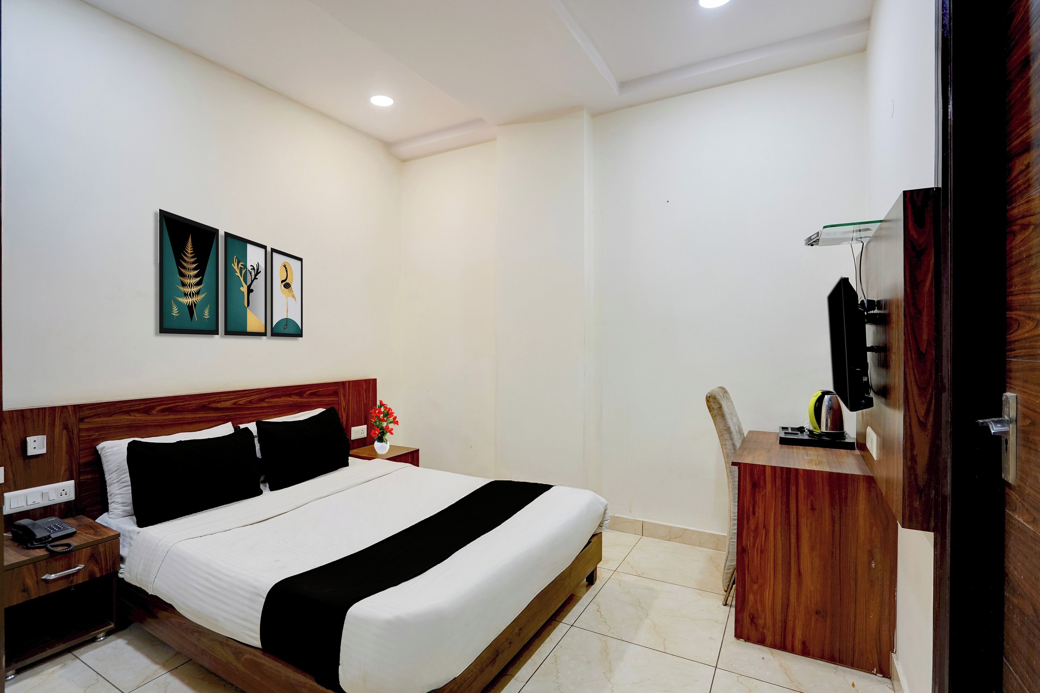 A hotel room with a bed, bedside table, a small desk with a chair, a telephone, and a wall-mounted TV.