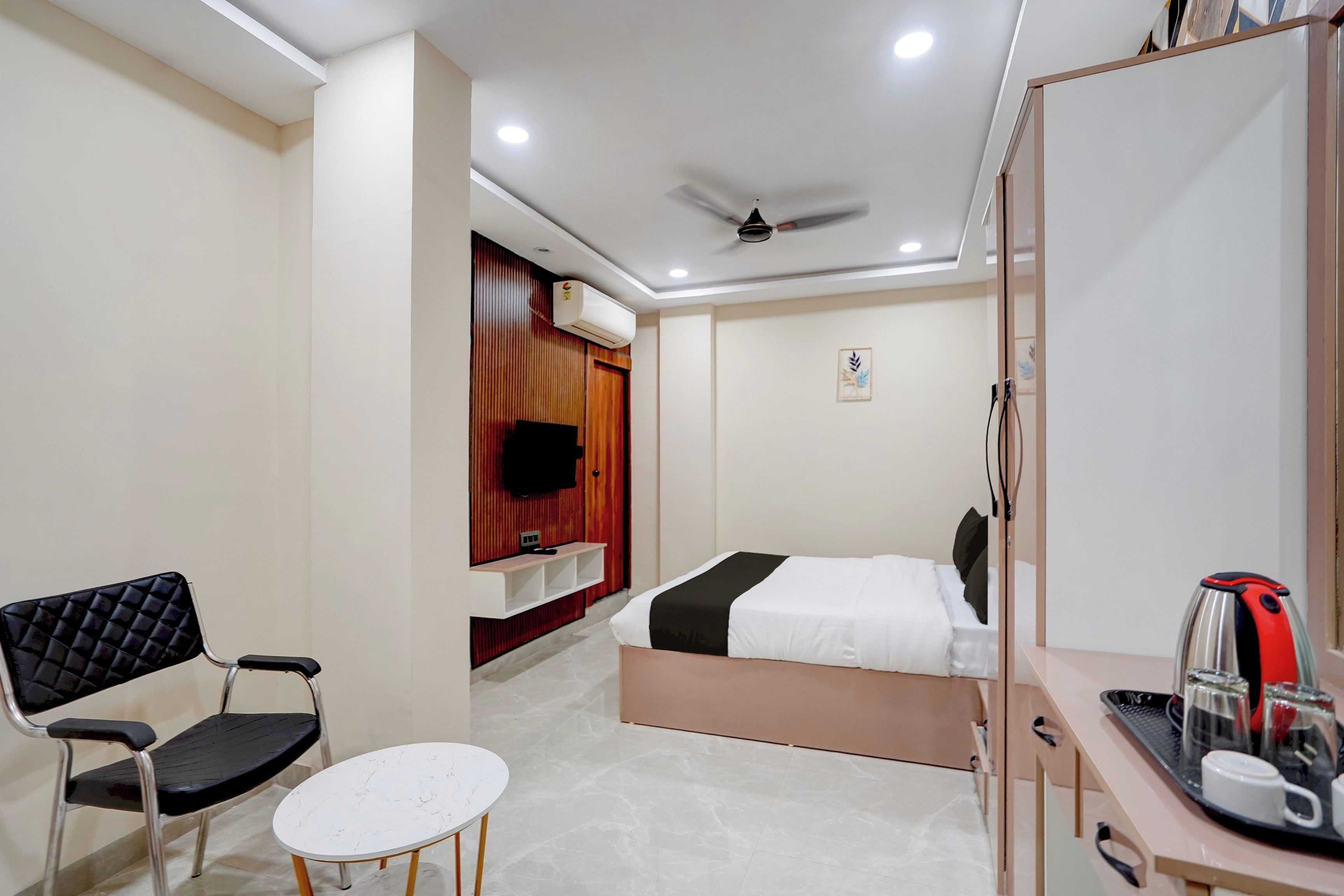 Habitación de hotel con cama, escritorio con tetera, silla, mesita pequeña, equipo de aire acondicionado y ventilador de techo.