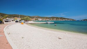 A pebble beach with clear turquoise water, a small boat docked, and a few people in the distance. - A3 - Modern apartment with garden and balcony with sea view, 80m from beach (Vinišće)