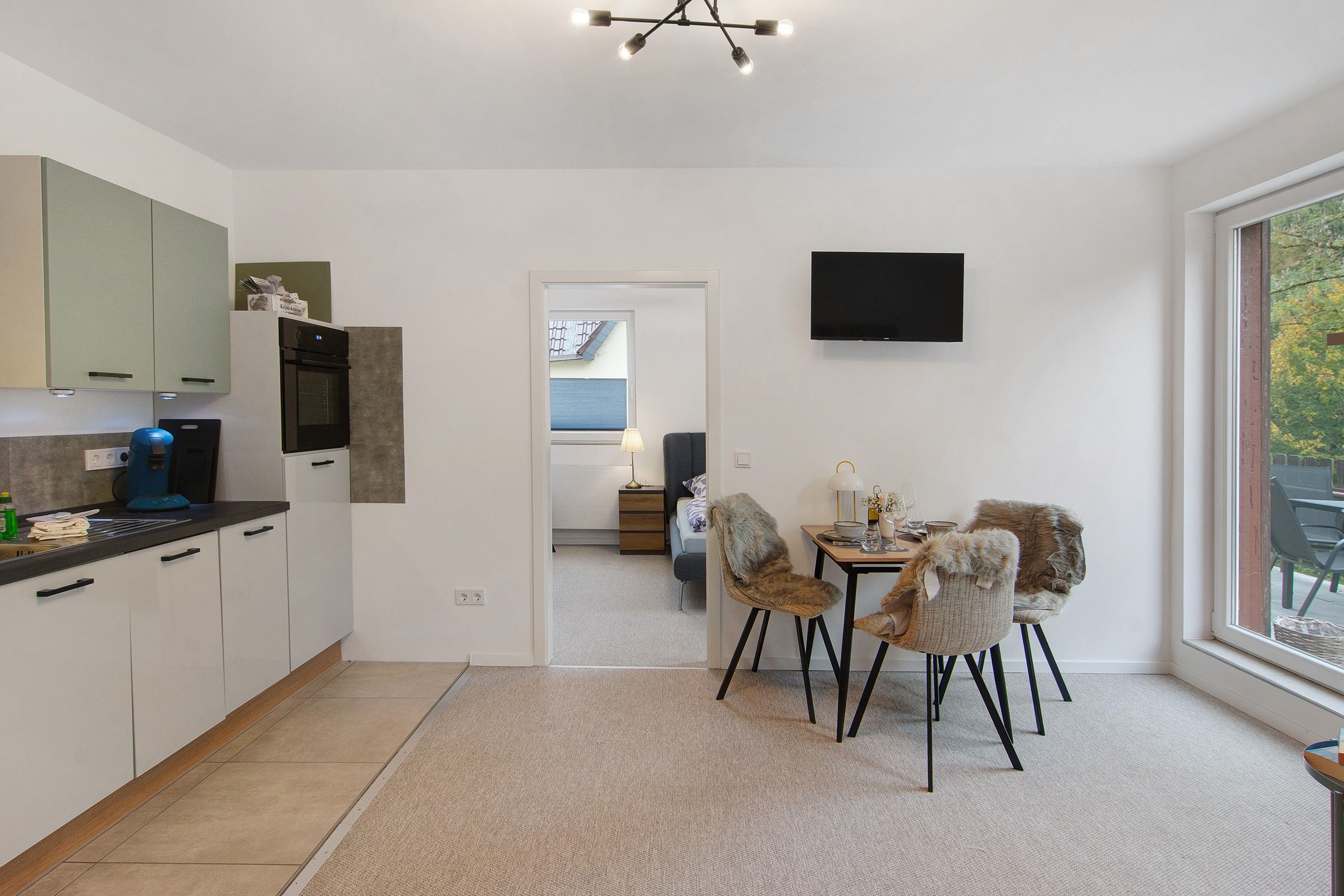 Eine moderne Küche mit Essbereich, einem an der Wand befestigten Flachbildfernseher und Blick ins Freie durch ein großes Fenster.