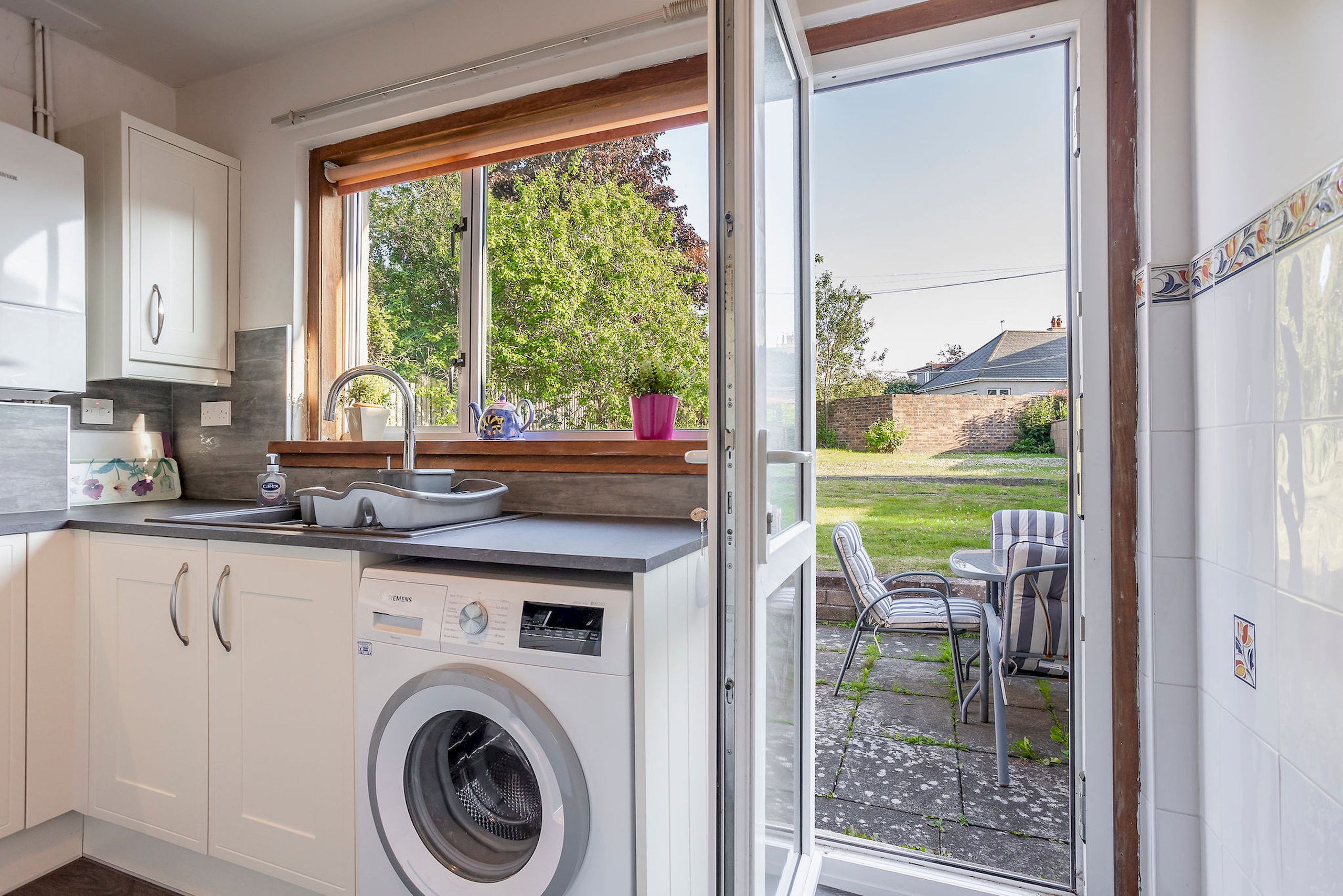 Une cuisine équipée d’un lave-linge intégré, une fenêtre donnant sur un jardin et un coin repas avec des sièges à l’extérieur.