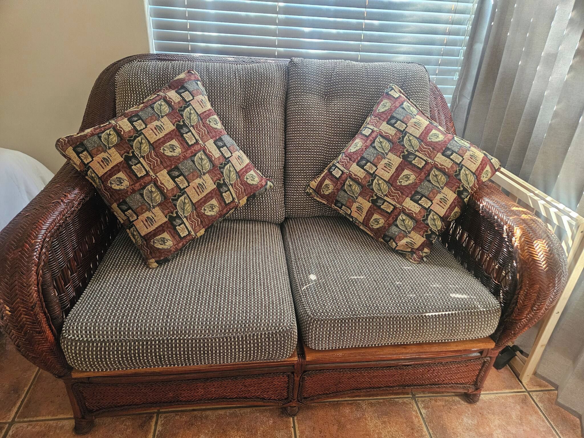 Un canapé en osier agrémenté de coussins à motifs, placé devant une fenêtre munie de stores.