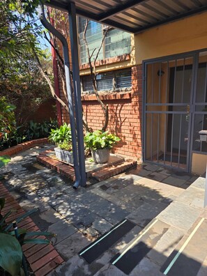 Un patio con pared de ladrillo, plantas en macetas, una reja metálica y suelo de baldosas.