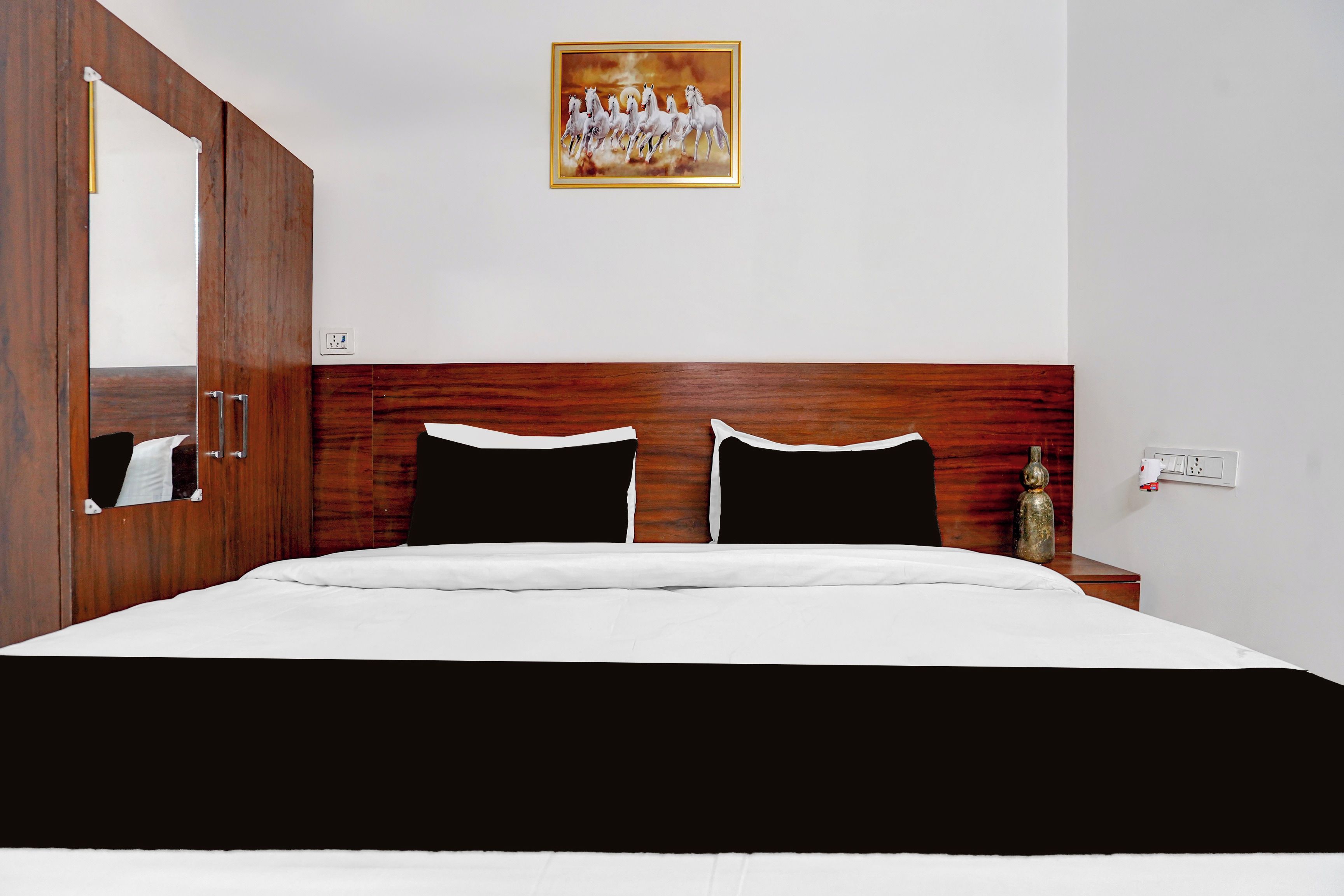 A bedroom with a wooden headboard, a bed with white bedding, a nightstand with a lamp, and a painting of horses on the wall.