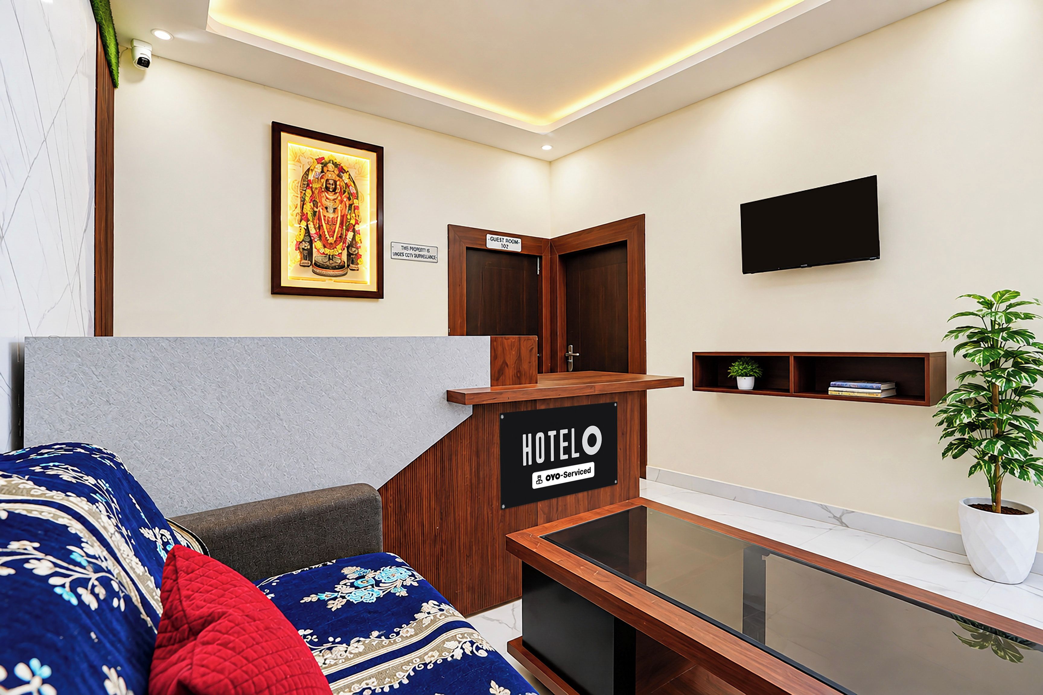 A hotel room with a sofa, a framed picture on the wall, a sign indicating a guest room, a flat-screen TV mounted on the wall, and a potted plant.