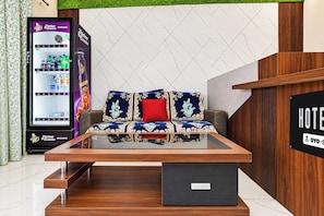 A hotel reception area with a sofa, a laptop on a stand, a refrigerator stocked with beverages, and a sign indicating the hotel name. - Hotel O Ayodhya Junction Road (Faizabad)