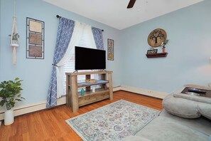A living room with a flat-screen TV on a wooden stand, a grey sofa, a patterned rug, and a wall-mounted shelf with decorative items. - Graceful Stays: Highlands 1BR near Dining – Unit 11 (Louisville)