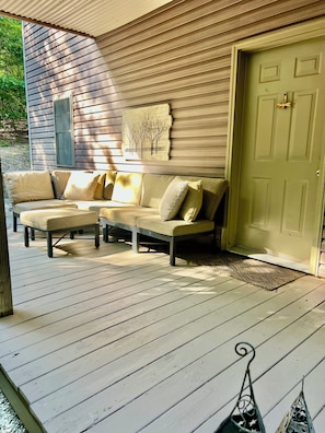 A wooden deck with a beige sofa and a matching bench, a door with a doormat, and a wall-mounted artwork. - Peaceful Laurel Lake Retreat with Private Waterfall Trail (Corbin)