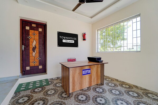 A reception desk labeled 'RECEPTION' with a fire extinguisher mounted on the wall, a door with a number sign, and a window with a view of greenery. - Townhouse Mattuthavani (Madurai)