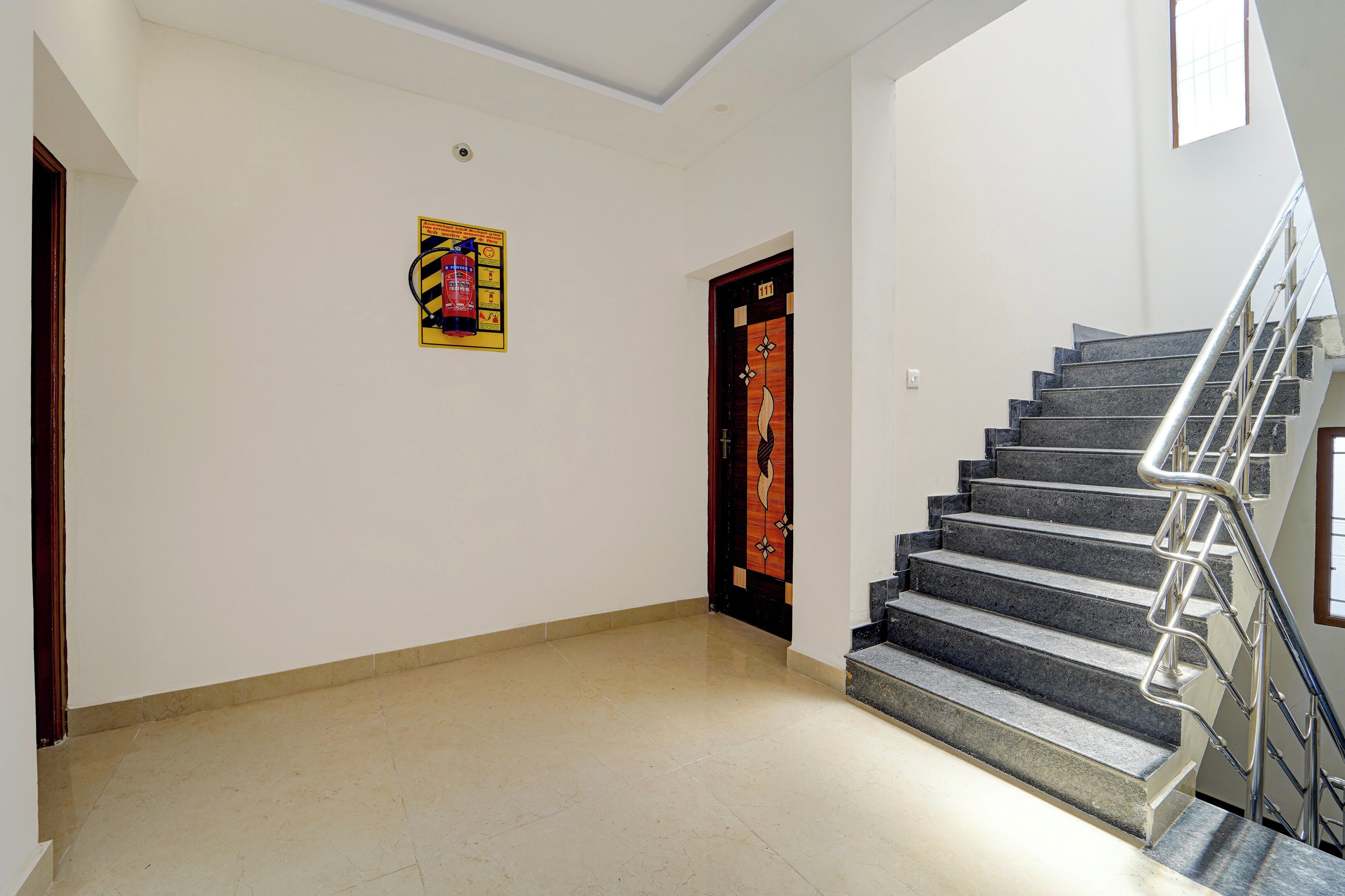 Un pasillo con piso de baldosas, un extintor de incendios en la pared, una puerta con diseño decorativo y una escalera con barandales de metal.