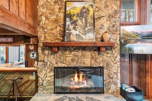 A stone fireplace with a fire, a wooden bar area, and a framed picture of a cabin on a mountain. - Silverthorne Mountain Retreat Hot Tub Sauna Near Keystone Breck and Copper (Silverthorne)