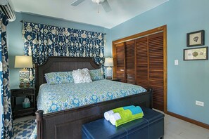 Ein Schlafzimmer mit einem Bett, Nachttischen, einer Bank, einem Deckenventilator und einem Fenster mit Holzjalousien.