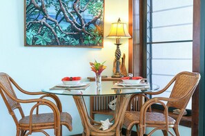 Un comedor con mesa de vidrio, sillas de mimbre, un cuadro de un árbol, una lámpara y una ventana con persianas.