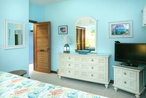 A bedroom with a bed, dresser, television, and a door leading to another room. - Lavender Hill - Mermaid’s Tale #10E – Renovated 1-Bedroom Condo with Cruz Bay Harbor Views (St John)