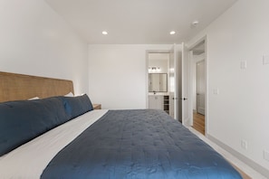 Quarto com cama, cabeceira de madeira, mesa de cabeceira e banheiro visível por uma porta aberta.
