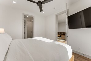 Ein modernes Schlafzimmer mit einem Bett, einem Deckenventilator, einem Fernseher und einem durch eine offene Tür einsehbarer Badezimmer.