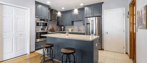 Una cocina moderna con armarios grises, electrodomésticos de acero inoxidable, una isla central con taburetes e iluminación colgante.