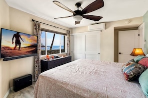 A bedroom with a bed, a ceiling fan, a TV on the wall, a window with curtains, and a door. - Kona Reef#A1: Sand & Surf Beachfront Corner, No Stairs (Kailua Kona)