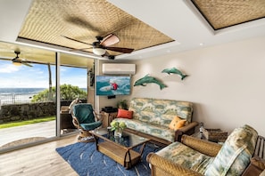 A spacious living area with a large window overlooking a scenic view, a ceiling fan, and a television displaying a surfer. - Kona Reef#A1: Sand & Surf Beachfront Corner, No Stairs (Kailua Kona)